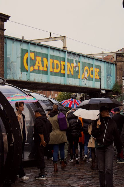 A crowded street scene beneath a weathered blue rail freight bridge with yellow lettering spelling 'CANDEN LOCK,' over a group of pedestrians holding umbrellas in various colours including a Union Jac