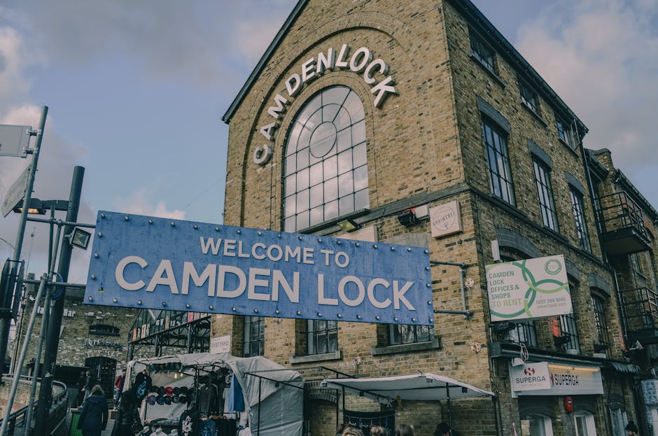 A crowded street scene beneath a weathered blue rail freight bridge with yellow lettering spelling 'CANDEN LOCK,' over a group of pedestrians holding umbrellas in various colours including a Union Jac