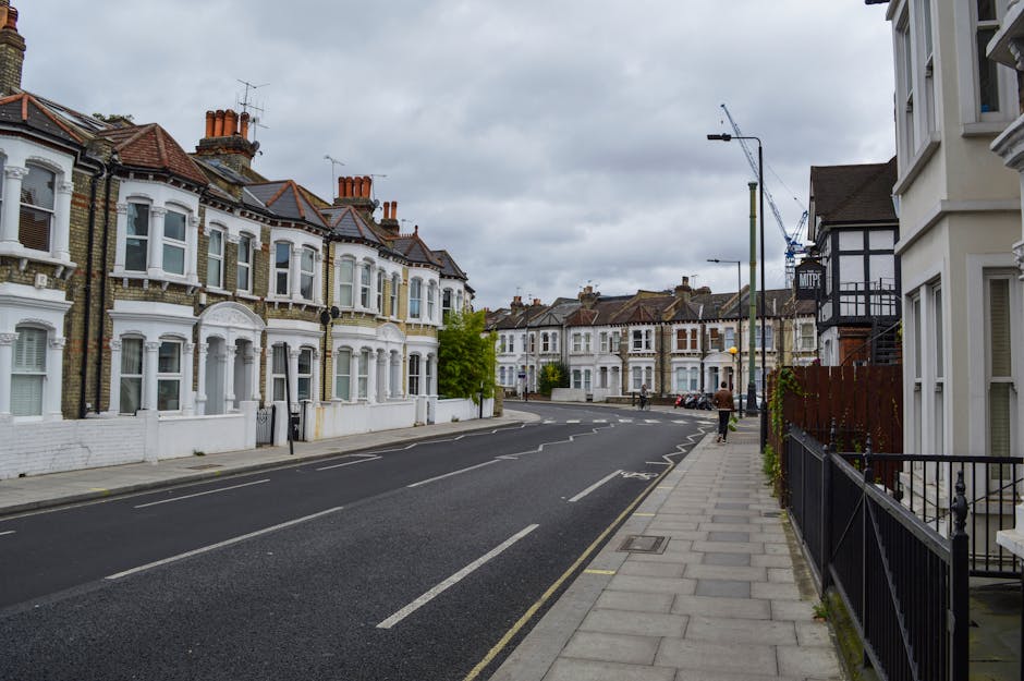A quiet residential street scene during daytime featuring a row of Victorian terraced houses with bay windows, brick facades, and chimney stacks. The pavement runs alongside the houses, with a black m