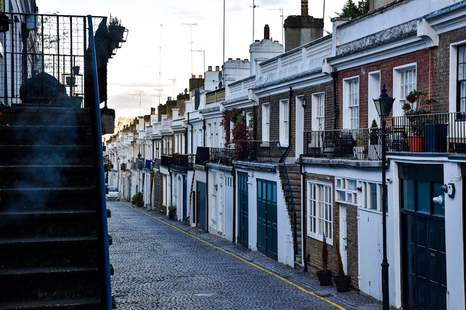 A quiet residential street scene during daytime featuring a row of Victorian terraced houses with bay windows, brick facades, and chimney stacks. The pavement runs alongside the houses, with a black m