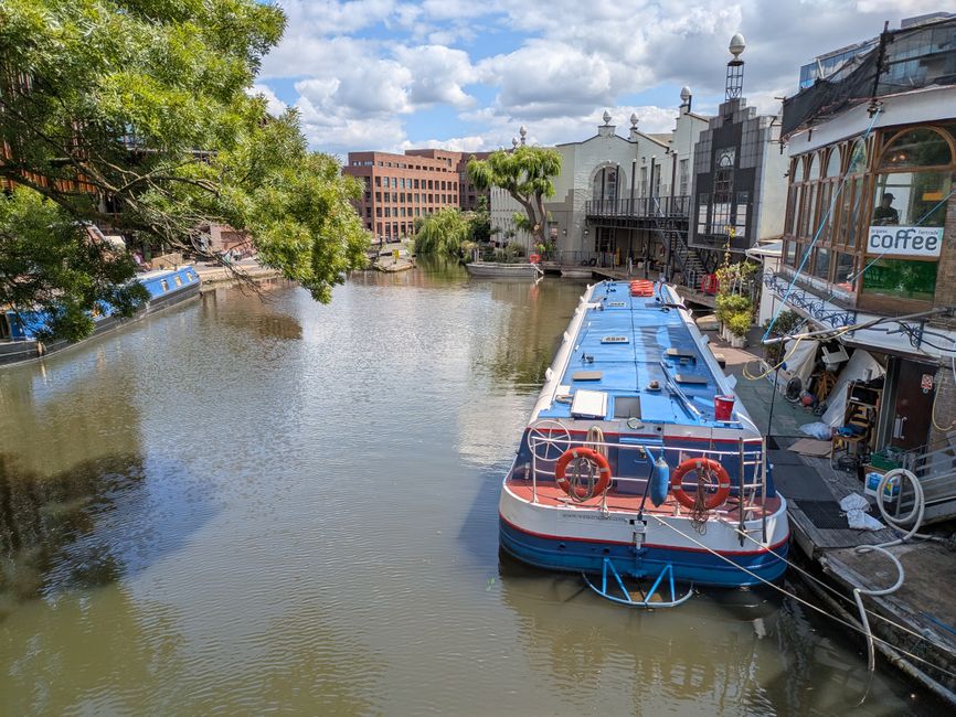 A daytime scene of a canal in Camden Town, with calm water reflecting the surrounding multi-storey residential buildings, some in yellow and white, lining the canal's edge. On the left, a houseboat is